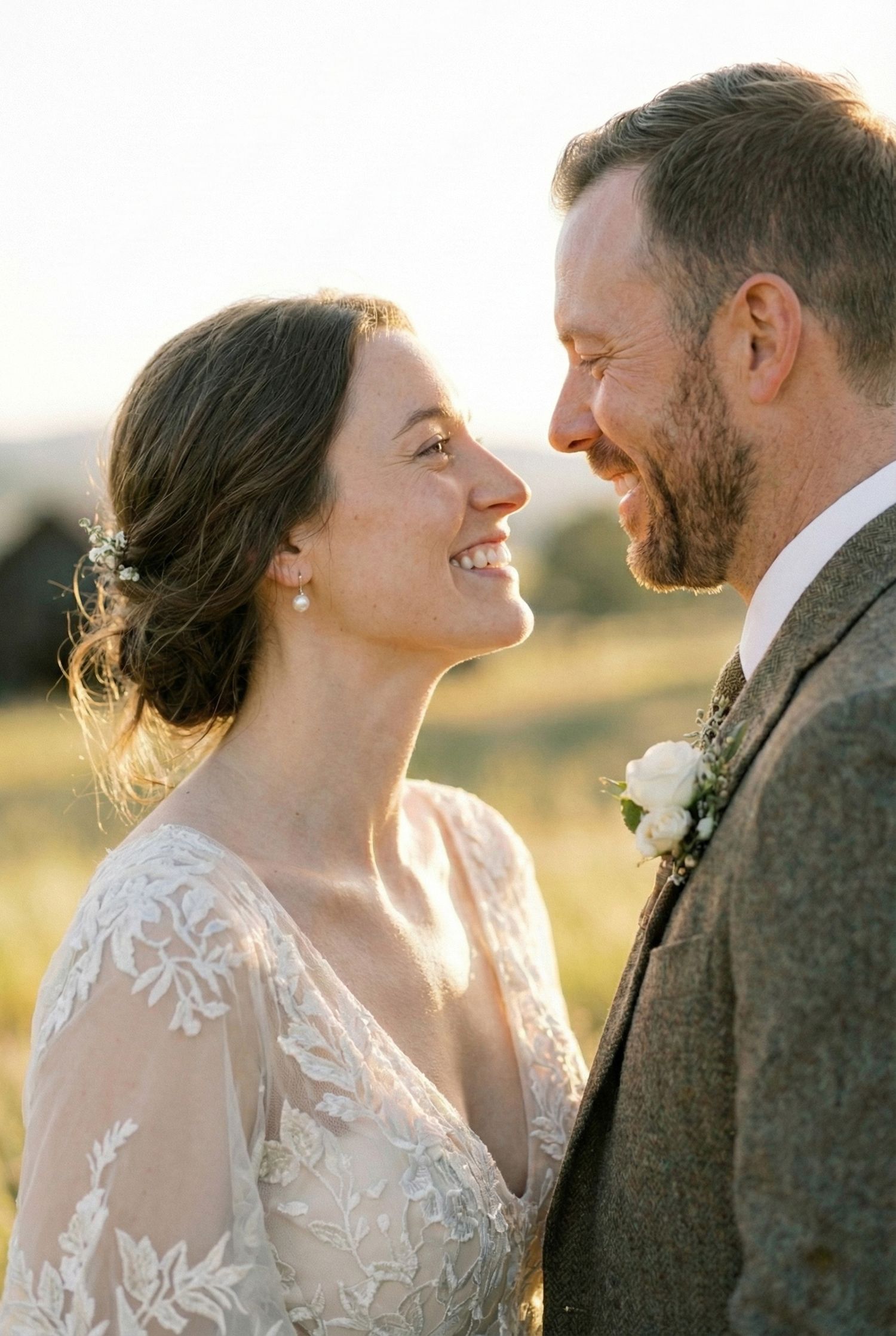 Smiling bride in lace gown and groom in tweed suit share a joyful moment outdoors in golden hour light.