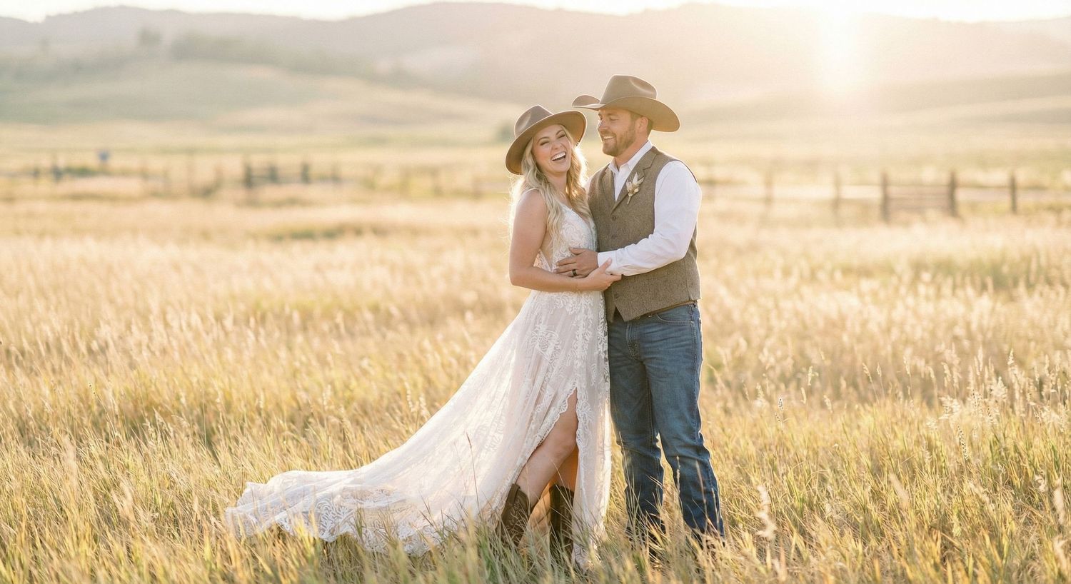 Couple in cowboy hats embracing in golden sunlit field, bride in lace gown, groom in vest and jeans, rustic wedding.
