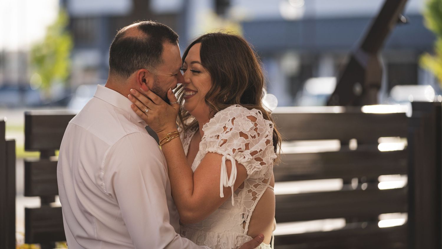 A couple shares a romantic moment, smiling and embracing outdoors near a wooden bench on a sunny day.