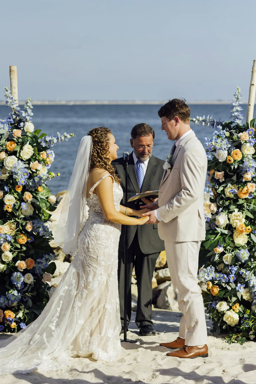 Bride and groom exchange vows at a beach wedding ceremony adorned with blue and orange floral arrangements.