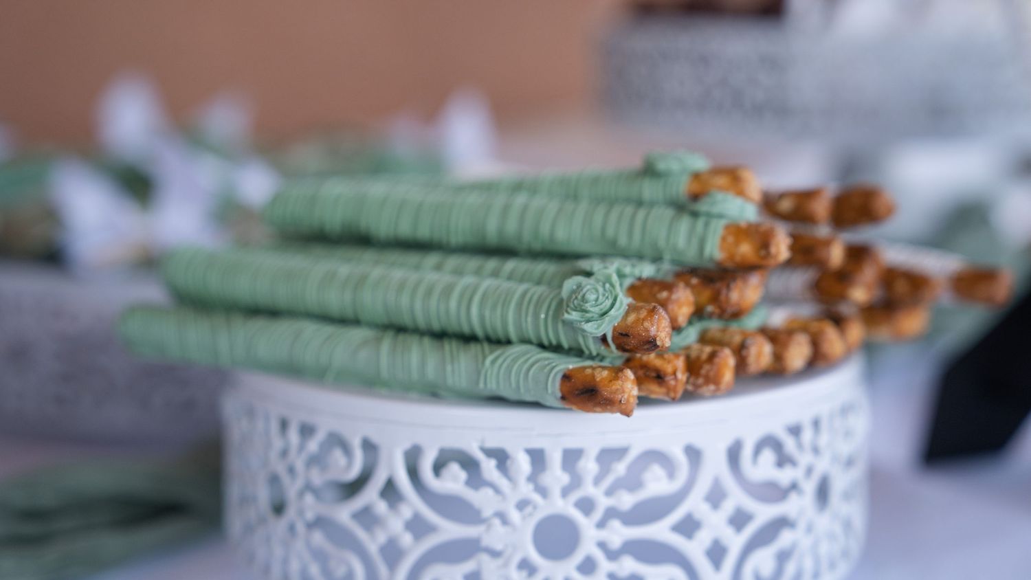 Green chocolate-dipped pretzel rods displayed on a decorative white lace cake stand at a dessert table.