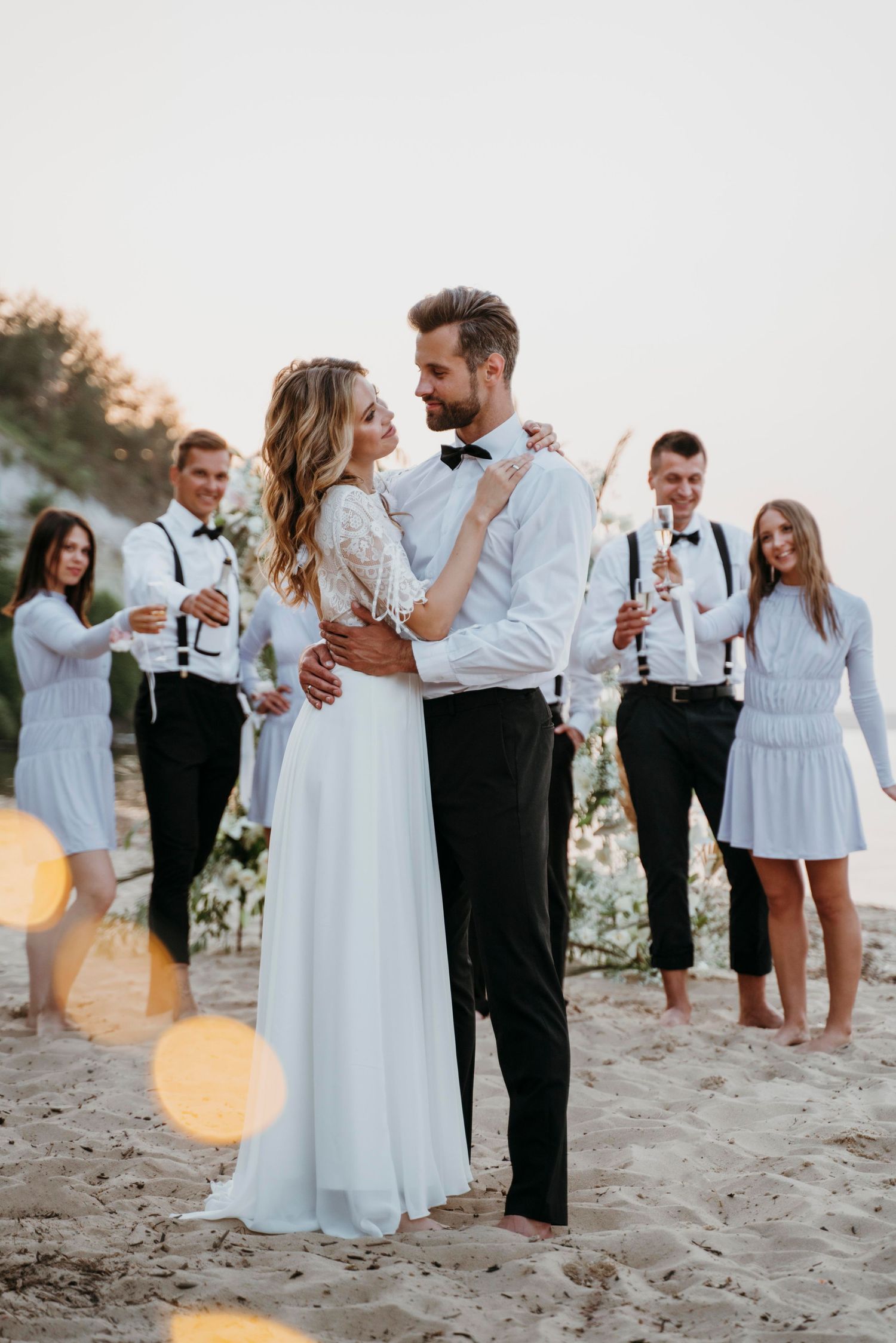Bride and groom embracing on beach wedding with bridal party in white attire holding champagne at sunset.