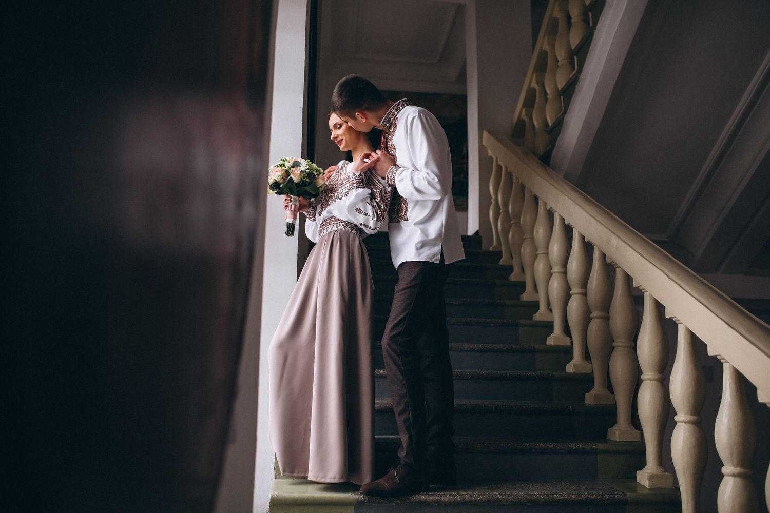 A romantic couple shares a tender moment on an elegant staircase, the bride holding a bouquet in a flowing gown.