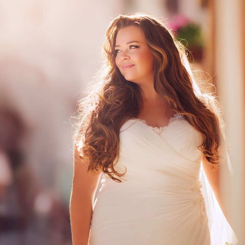A young woman in a white wedding dress with long wavy brown hair poses outdoors in soft sunlight.