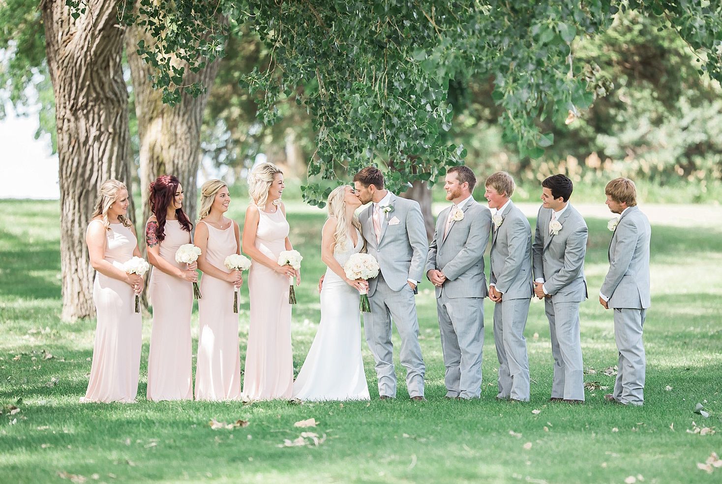 Wedding party poses outdoors under trees, bridesmaids in blush gowns and groomsmen in gray suits flanking the bride and groom.