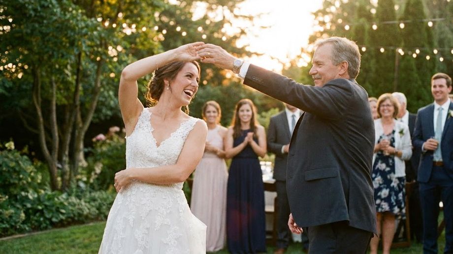 Bride in white lace gown twirling with older man in suit during outdoor wedding reception dance.