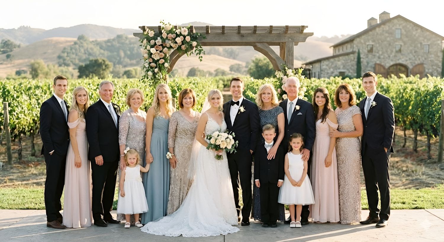Wedding party poses in vineyard with floral arch, bride in white gown, bridesmaids in blush and blue dresses.