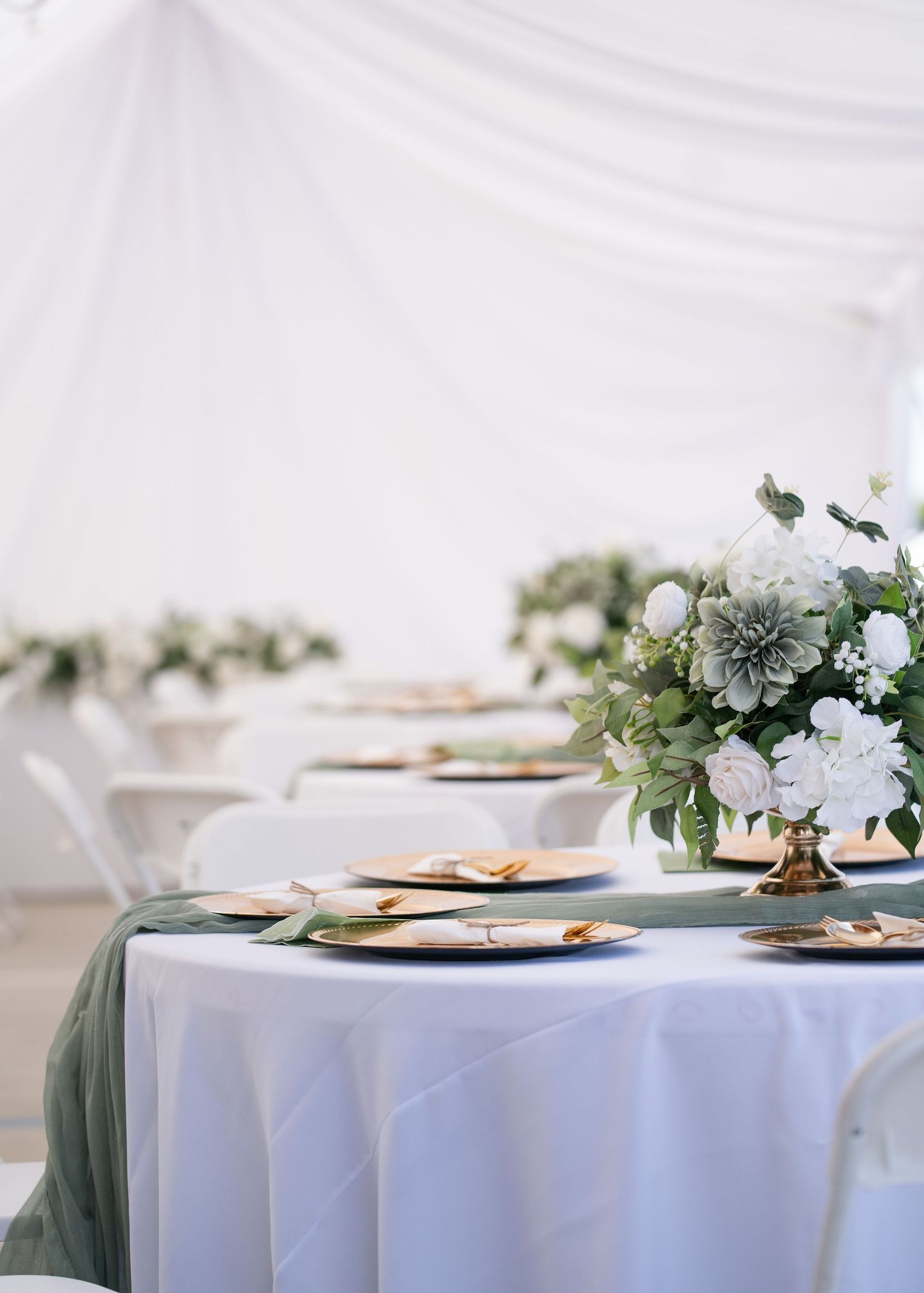 Elegant wedding reception table with white and sage green linens, gold chargers, and white floral centerpiece under a draped tent.