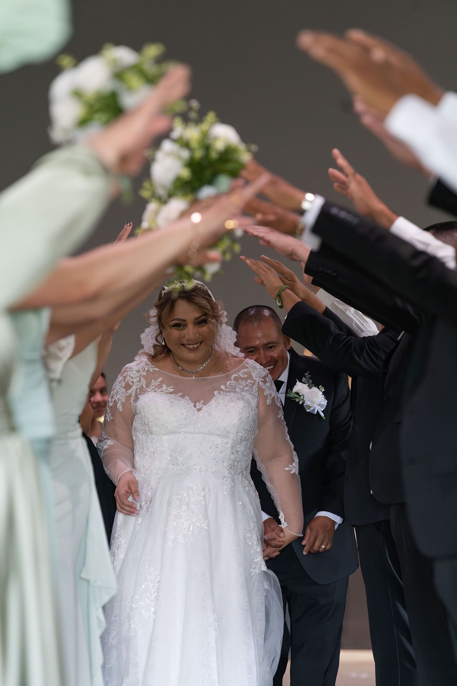 Bride in white lace gown walks with groom through arch of hands at wedding ceremony.