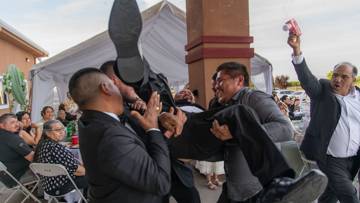 Men in suits scuffle at an outdoor event under a white tent, with onlookers watching the commotion.
