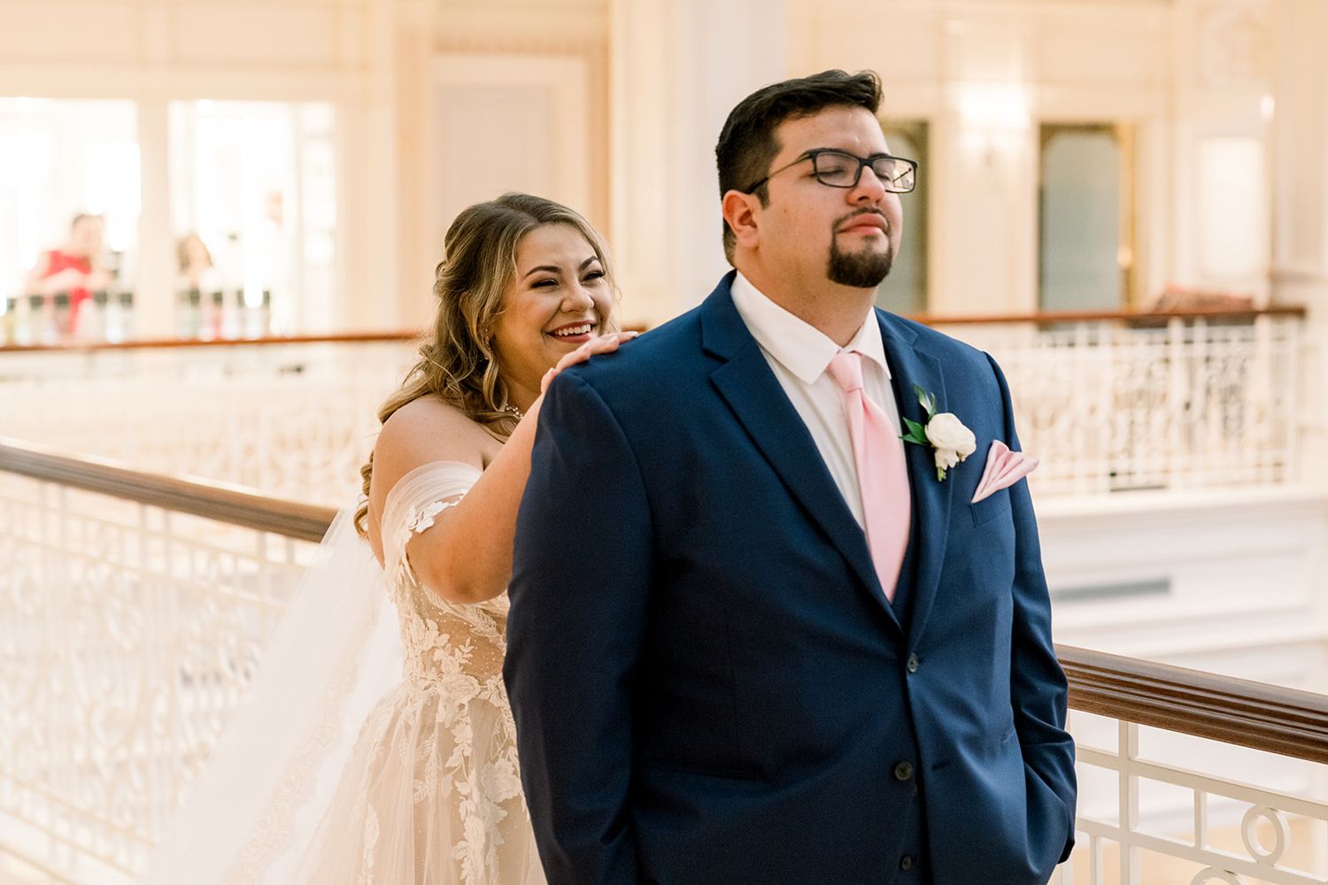 Bride in white gown smiles behind groom in navy suit with pink tie on elegant staircase at wedding venue.