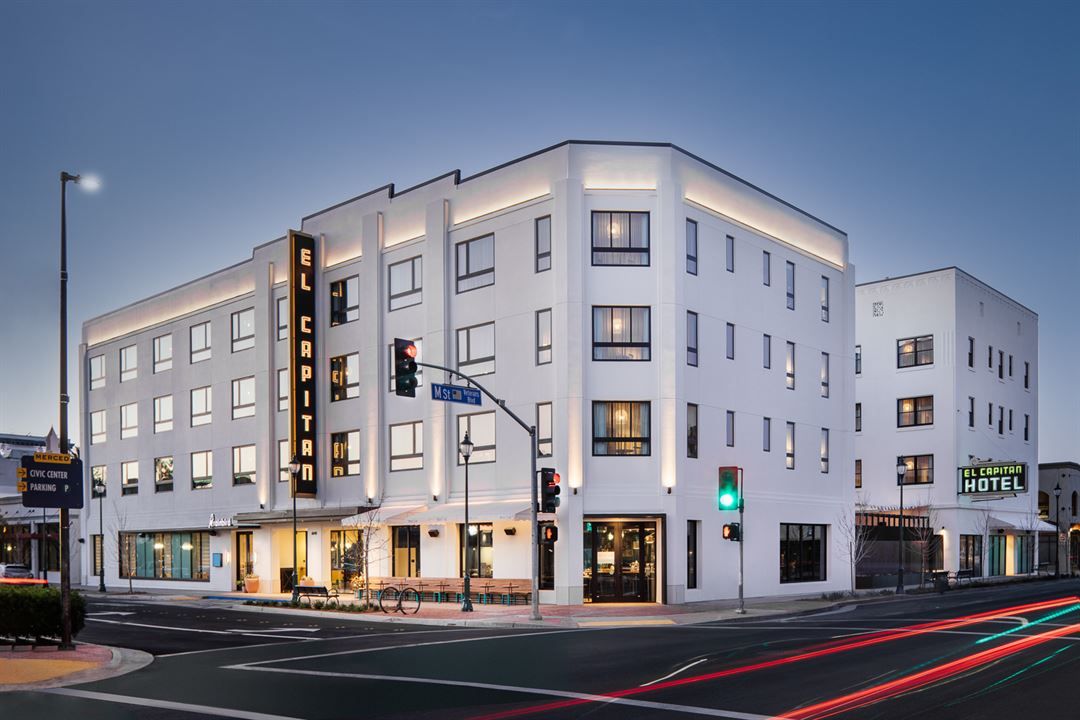 Modern white corner hotel building illuminated at dusk with street intersection and light trails in urban setting.