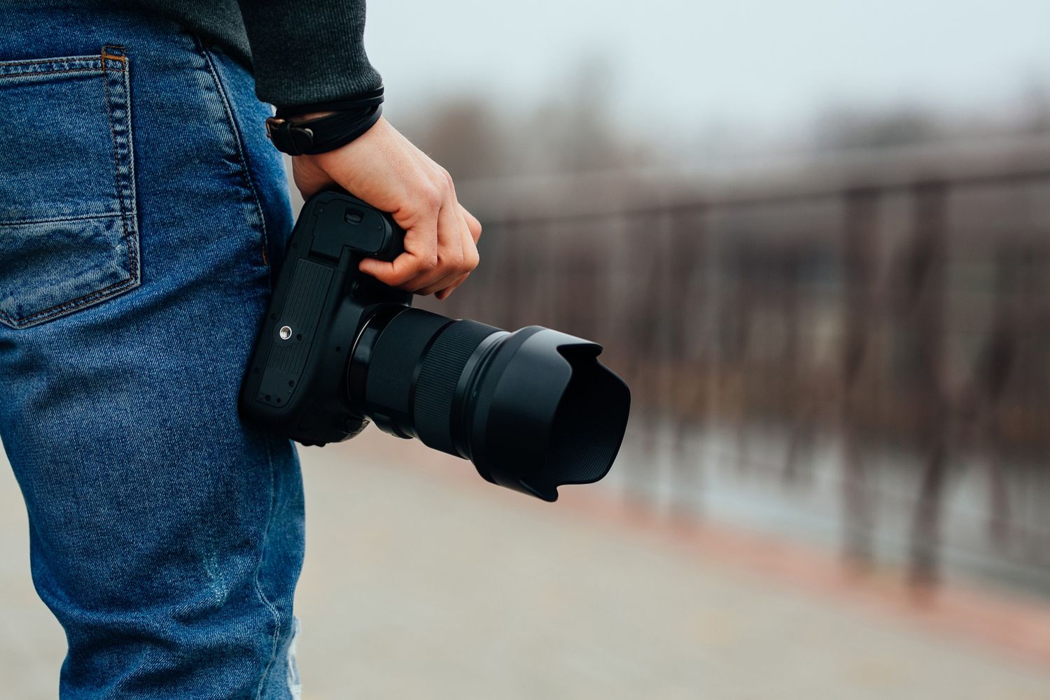 Photographer holding a professional DSLR camera with telephoto lens while standing outdoors in jeans. Merced Wedding Photographer