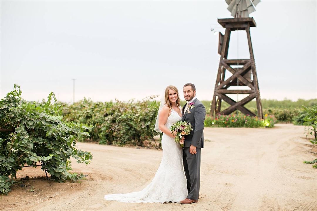Bride and groom pose on dirt path near rustic windmill tower surrounded by lush greenery at outdoor farm wedding.