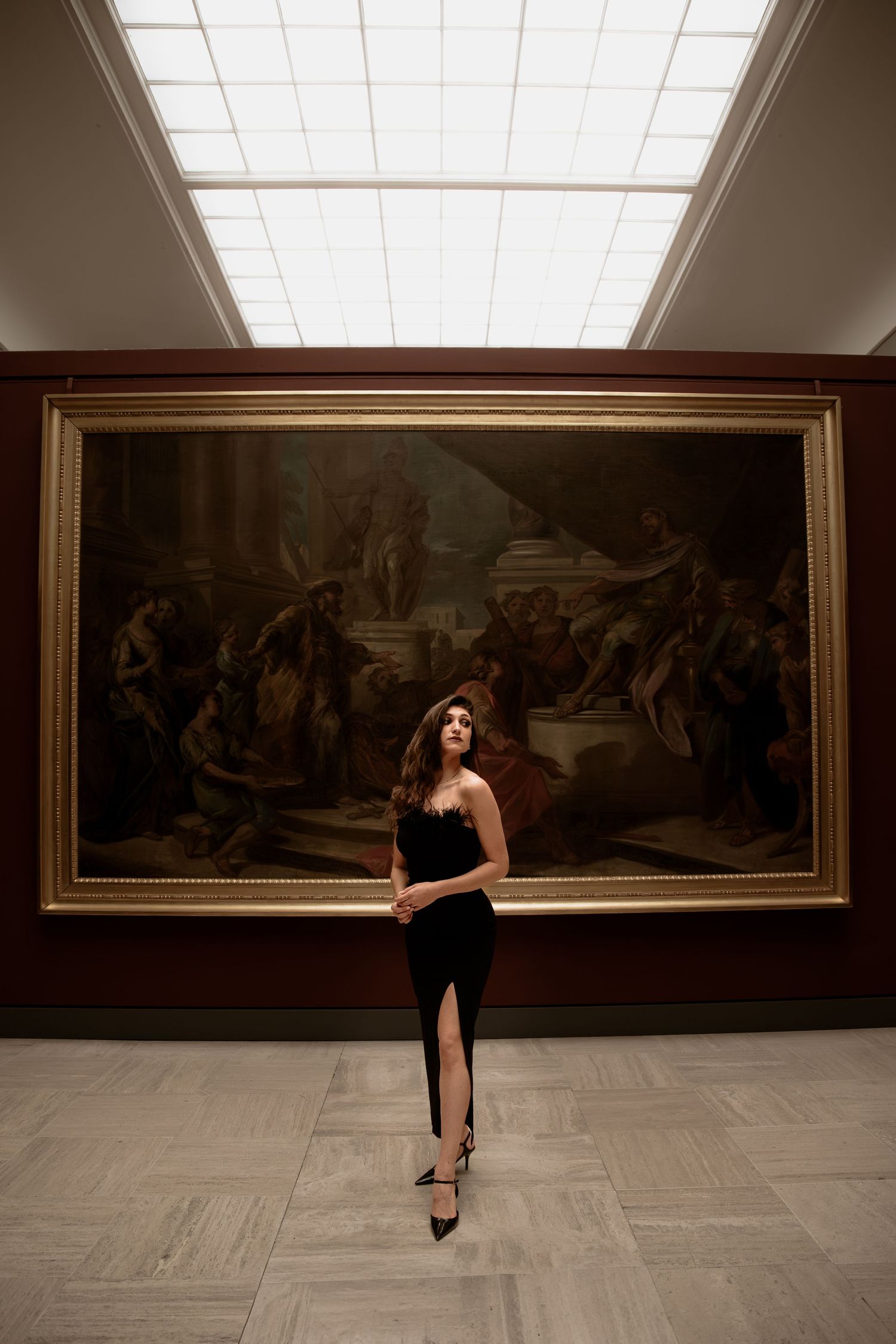 Woman in black dress poses before large classical painting in elegant museum gallery.