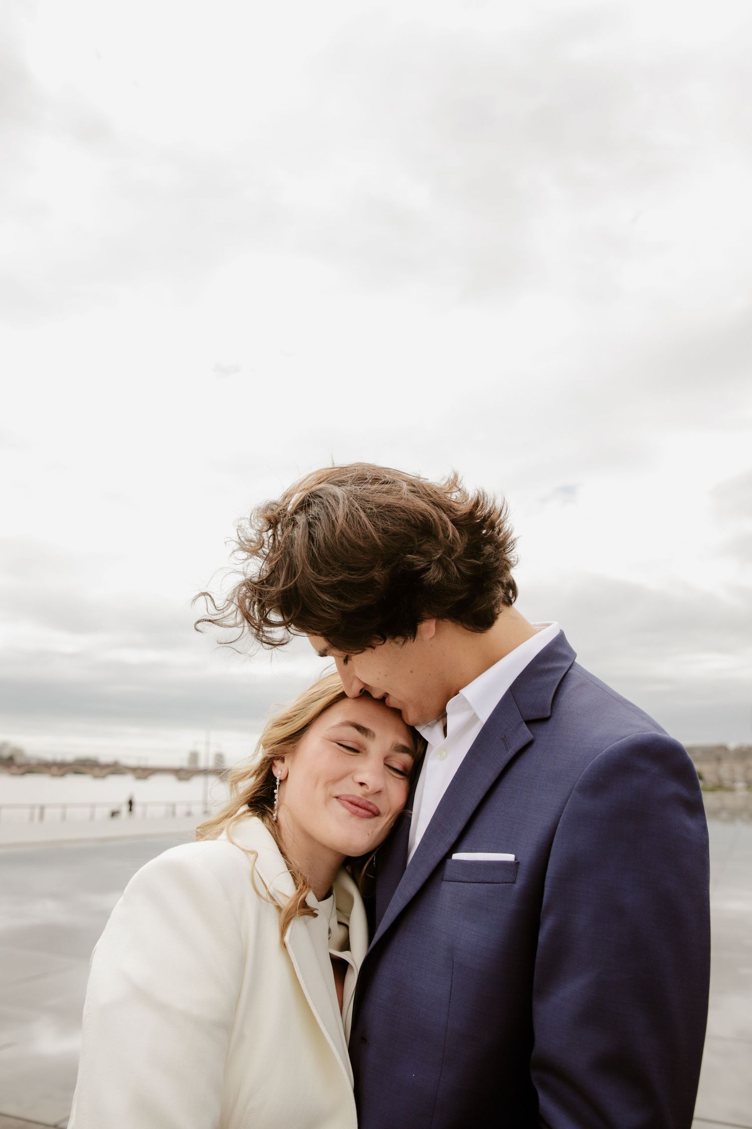 A couple shares a tender moment outdoors, man in navy suit kissing womans head as she smiles contentedly in white coat.