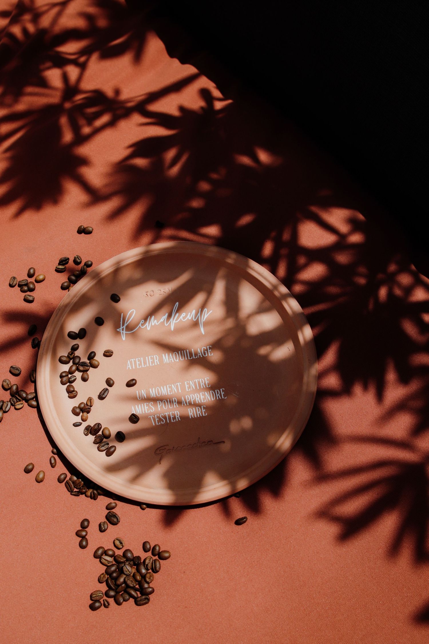 Overhead view of a round branded coffee tray with scattered roasted beans on a terracotta surface with leaf shadows.