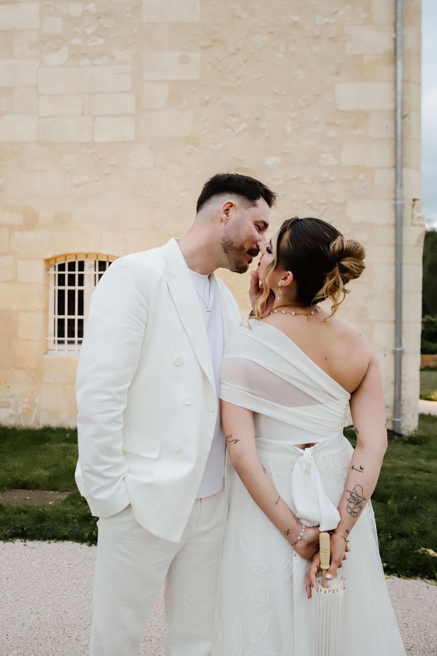 A couple in white wedding attire shares a romantic moment, kissing outdoors near a stone building.