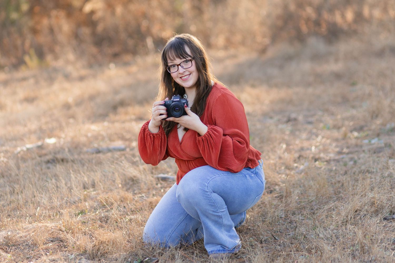 A young woman kneeling in field with her camera in photo taken by Little Buckaroo Portrait Co., an East Texas Newborn Photographer