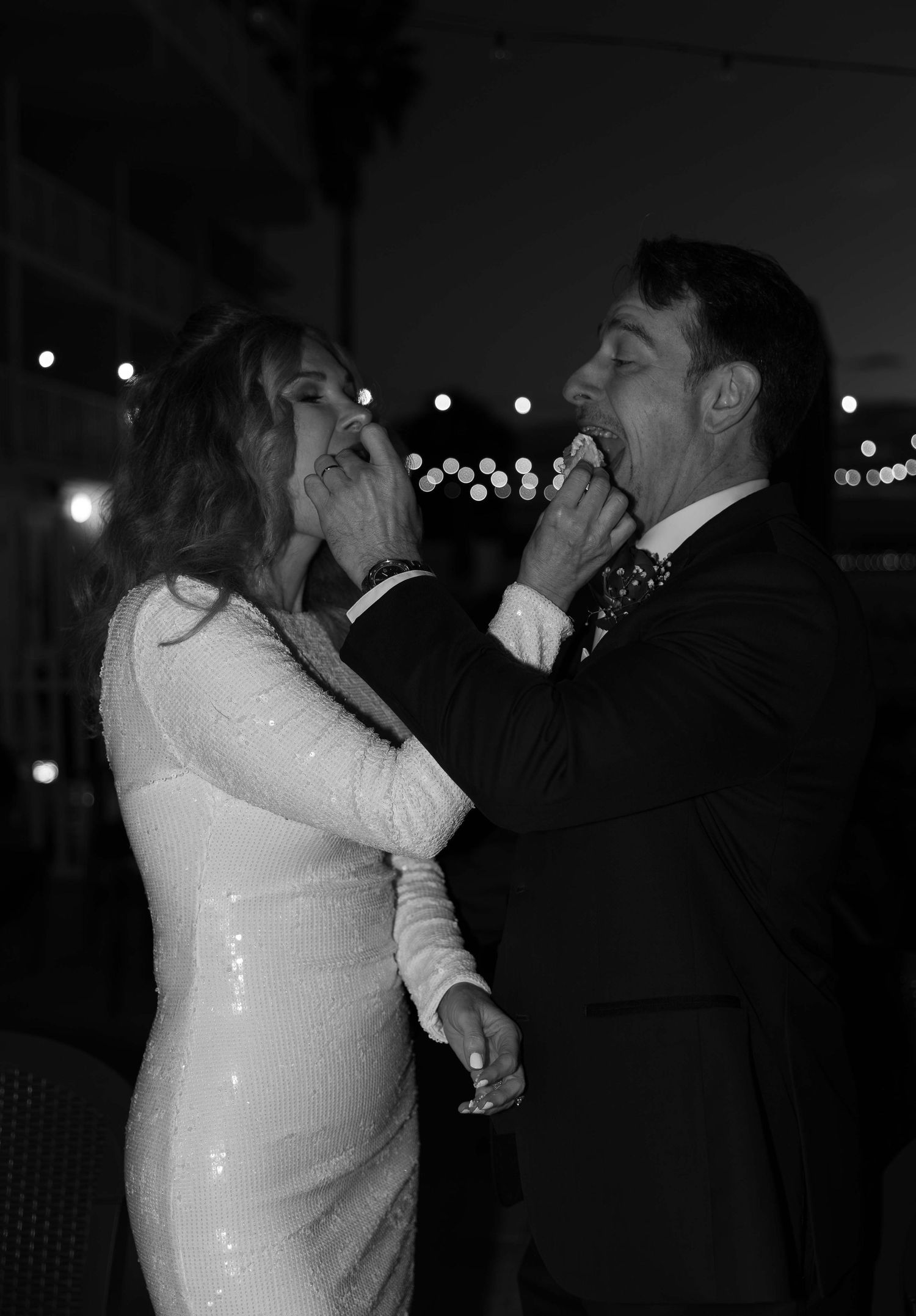 A playful wedding moment captured in black and white showing the couple squishing each other's cheeks with string lights in background.