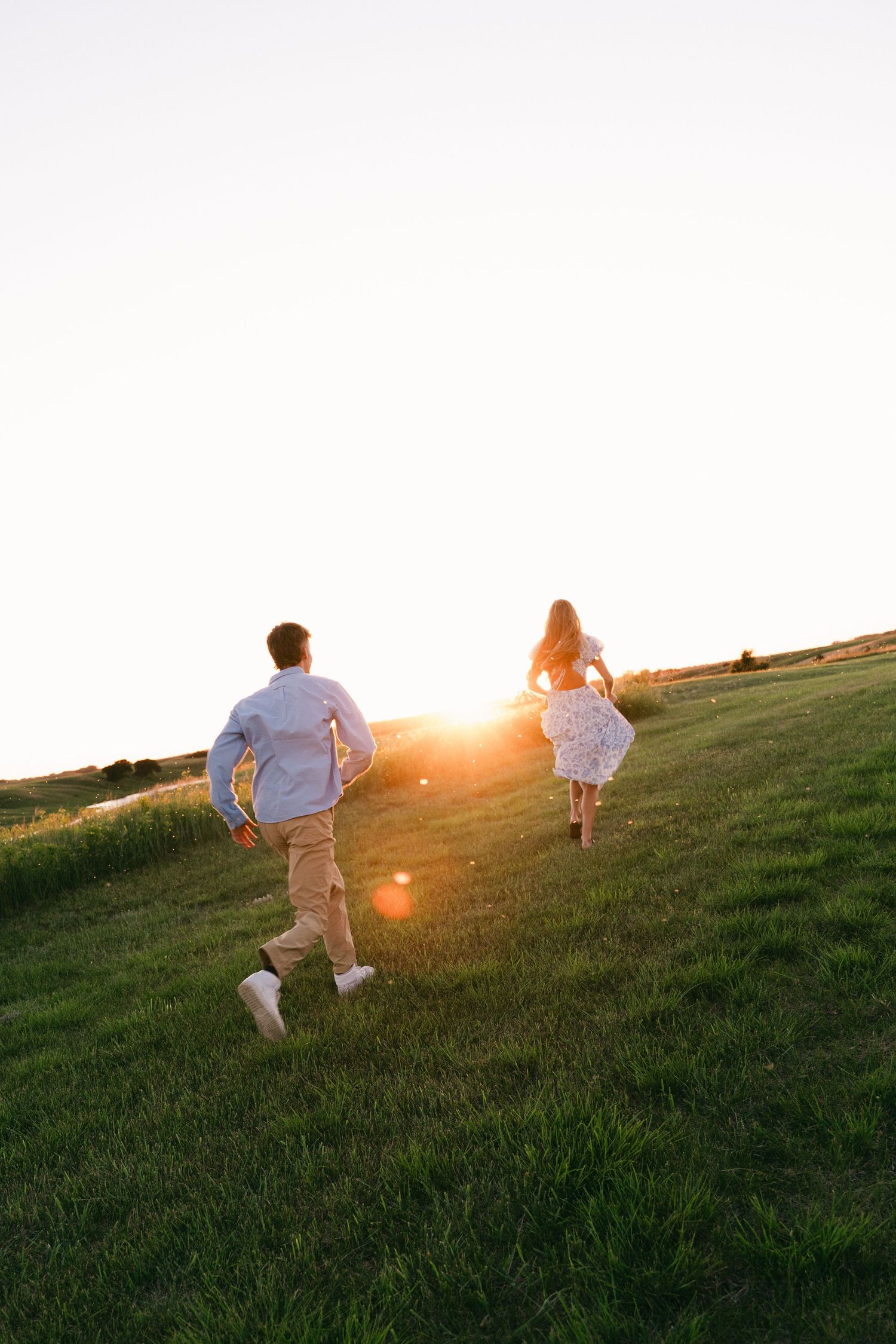 Couple running up a green grassy hill toward a golden sunset, viewed from behind in warm evening light.