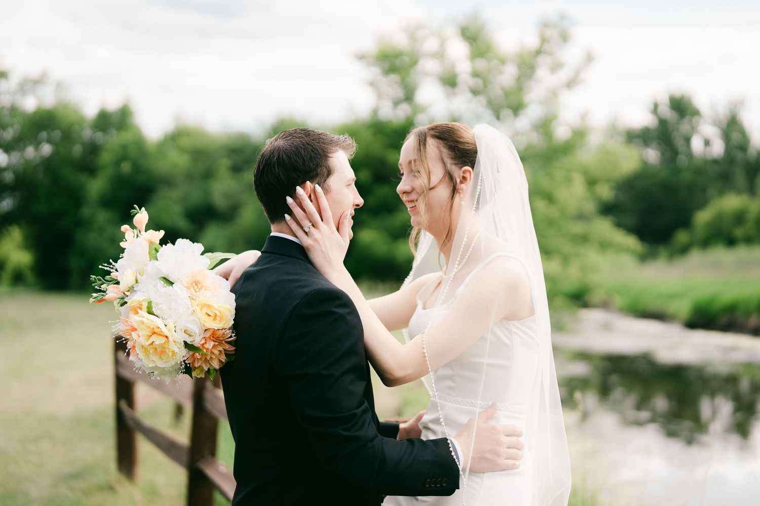 Bride and groom share a tender moment outdoors near a crick, with the bride holding a bouquet of white and yellow flowers.