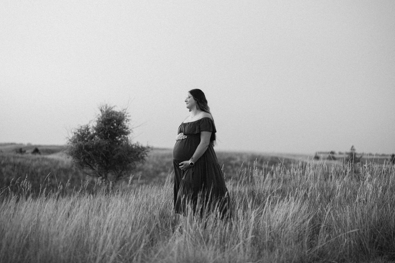 Pregnant woman in off-shoulder gown standing in tall grass field, black and white maternity portrait.