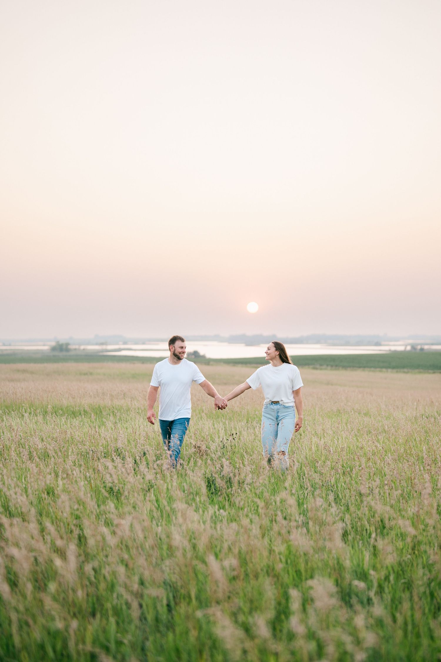 Couple in white t-shirts and jeans holding hands walking through a grassy field at sunset near a lake.