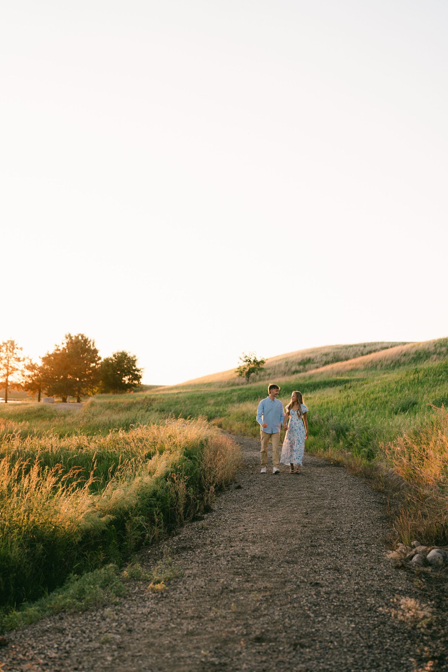 Couple holding hands walking on a dirt path through golden sunlit fields at sunset.