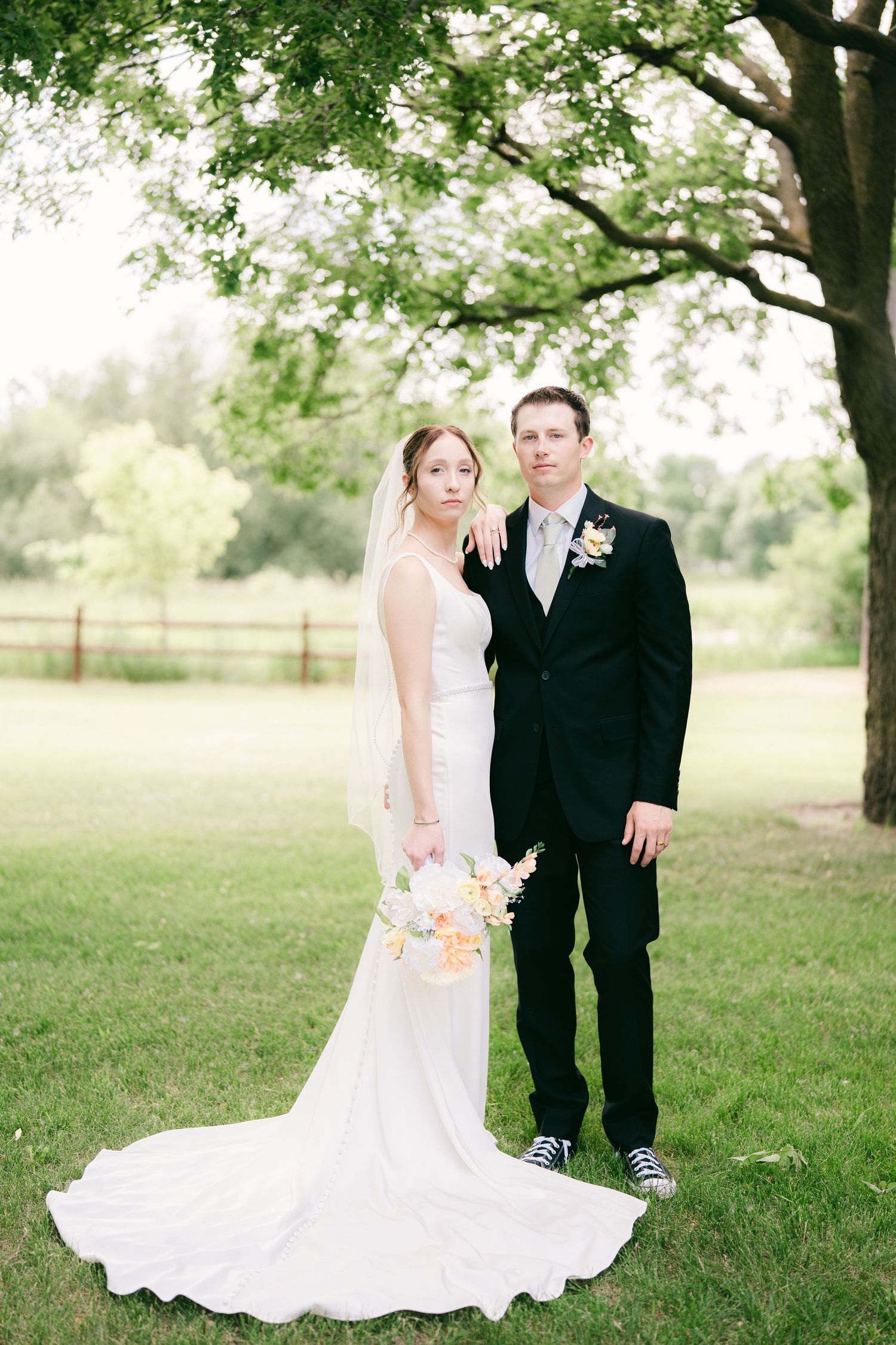 Bride in white gown with veil and groom in black suit stand together on green lawn under a tree.