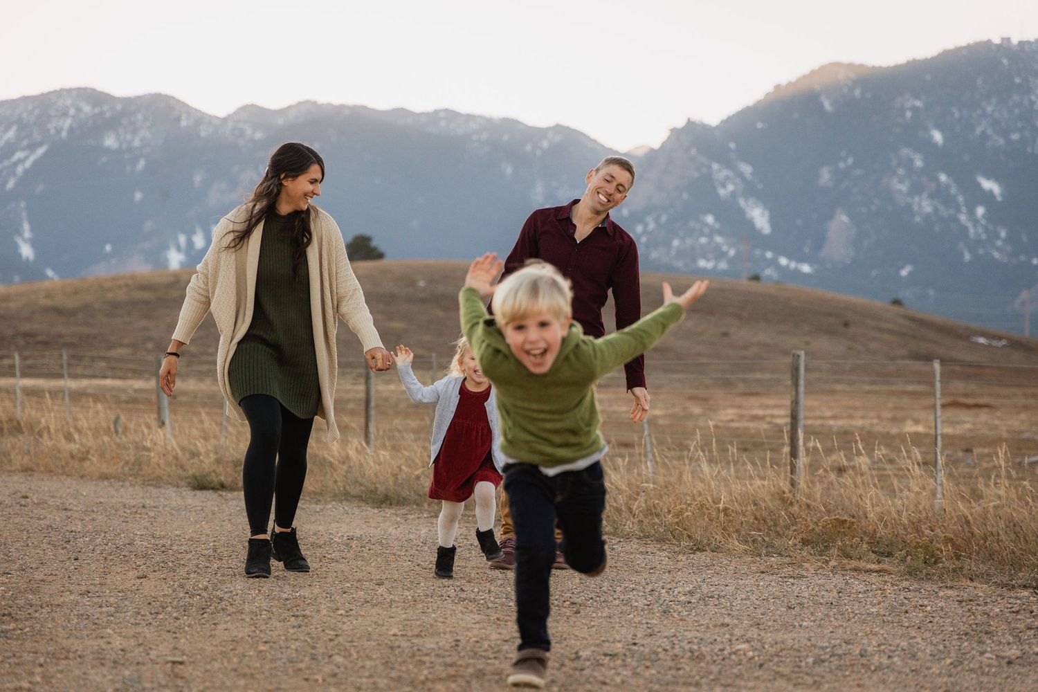 A couple holding hands and running playfully across a grassy meadow with mountains in the background.