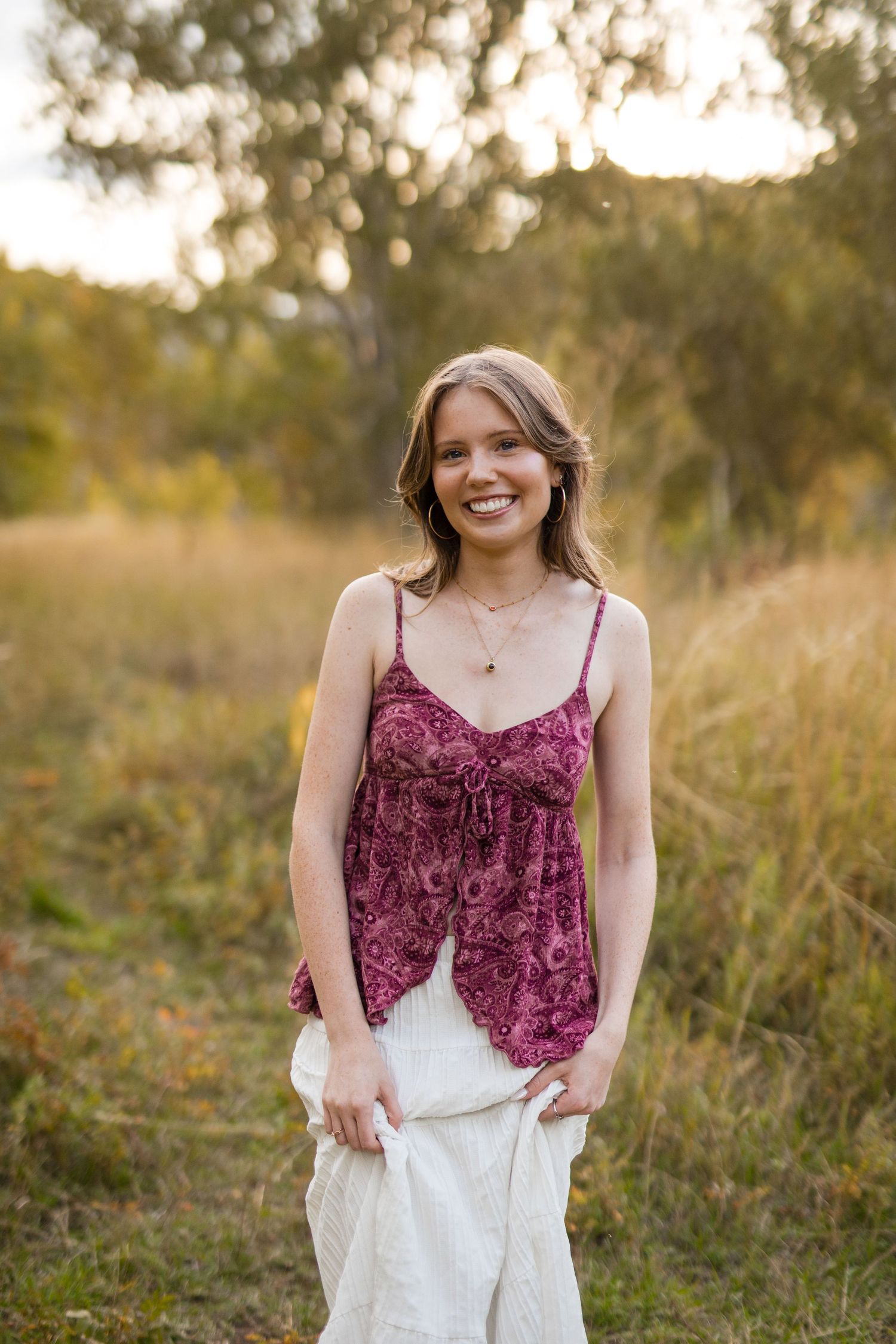 A high schooler in a burgundy velvet camisole and white skirt stands in a sunlit meadow during golden hour in Golden, Colorado