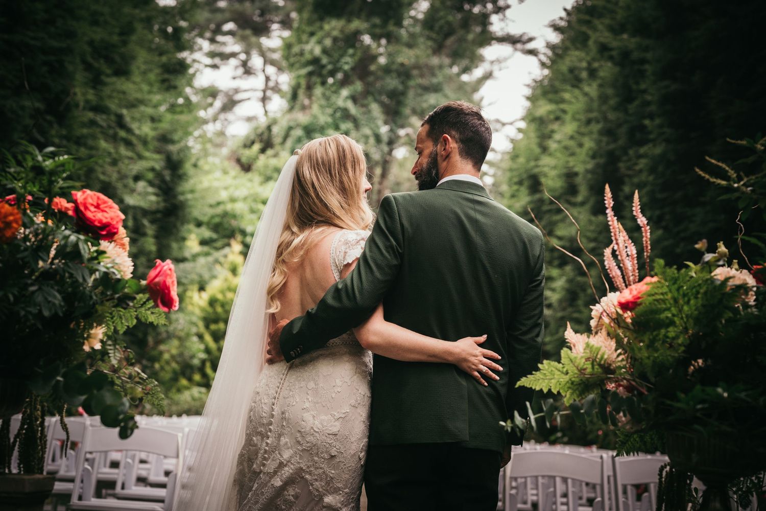 Bride and groom embracing outdoors at a garden wedding ceremony surrounded by lush greenery and colorful floral arrangements.