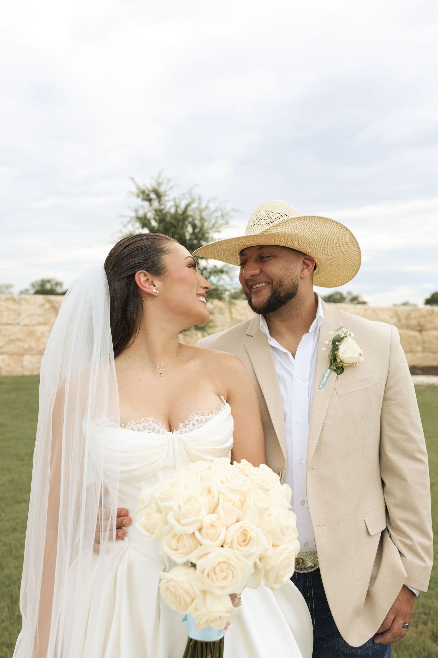 Newlyweds share intimate moments while holding cream-colored rose bouquet against cloudy sky.