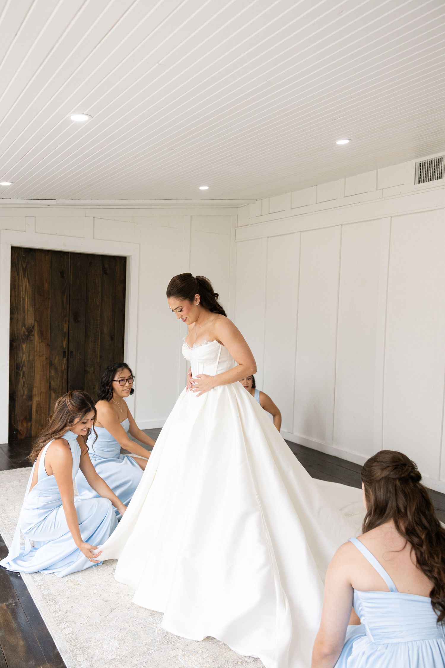 Bridesmaids in pale blue dresses help the bride with her full wedding gown in a white room.
