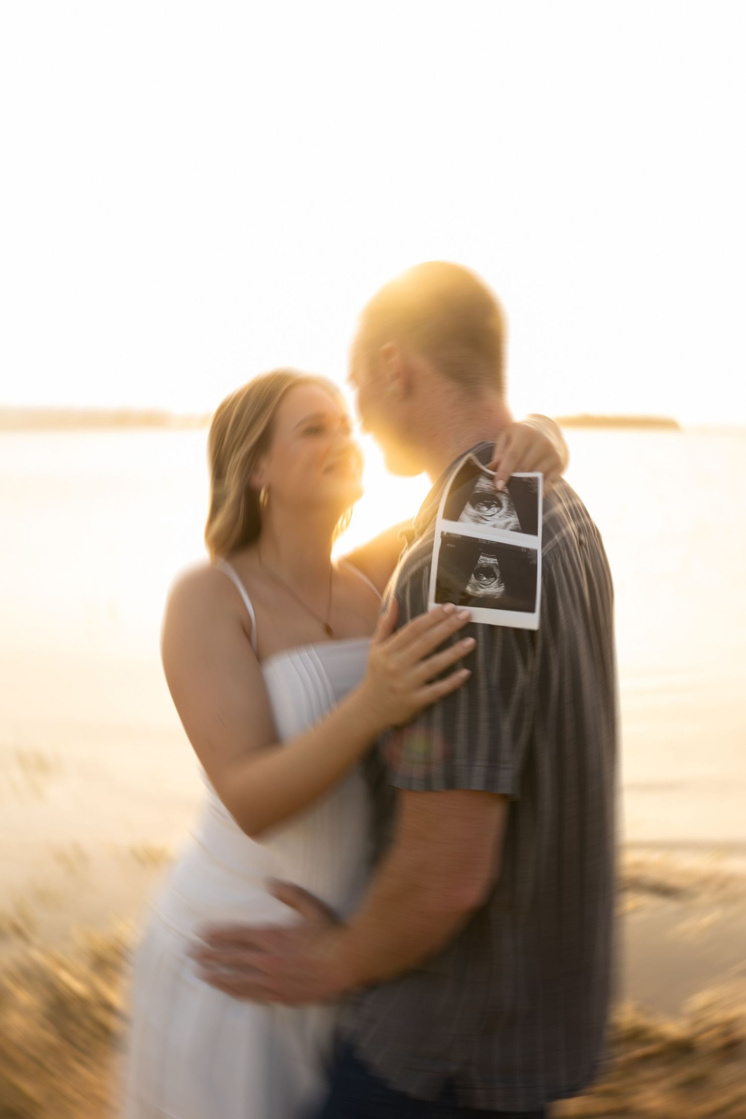 Couple embracing on beach with ultrasound photos at sunset.