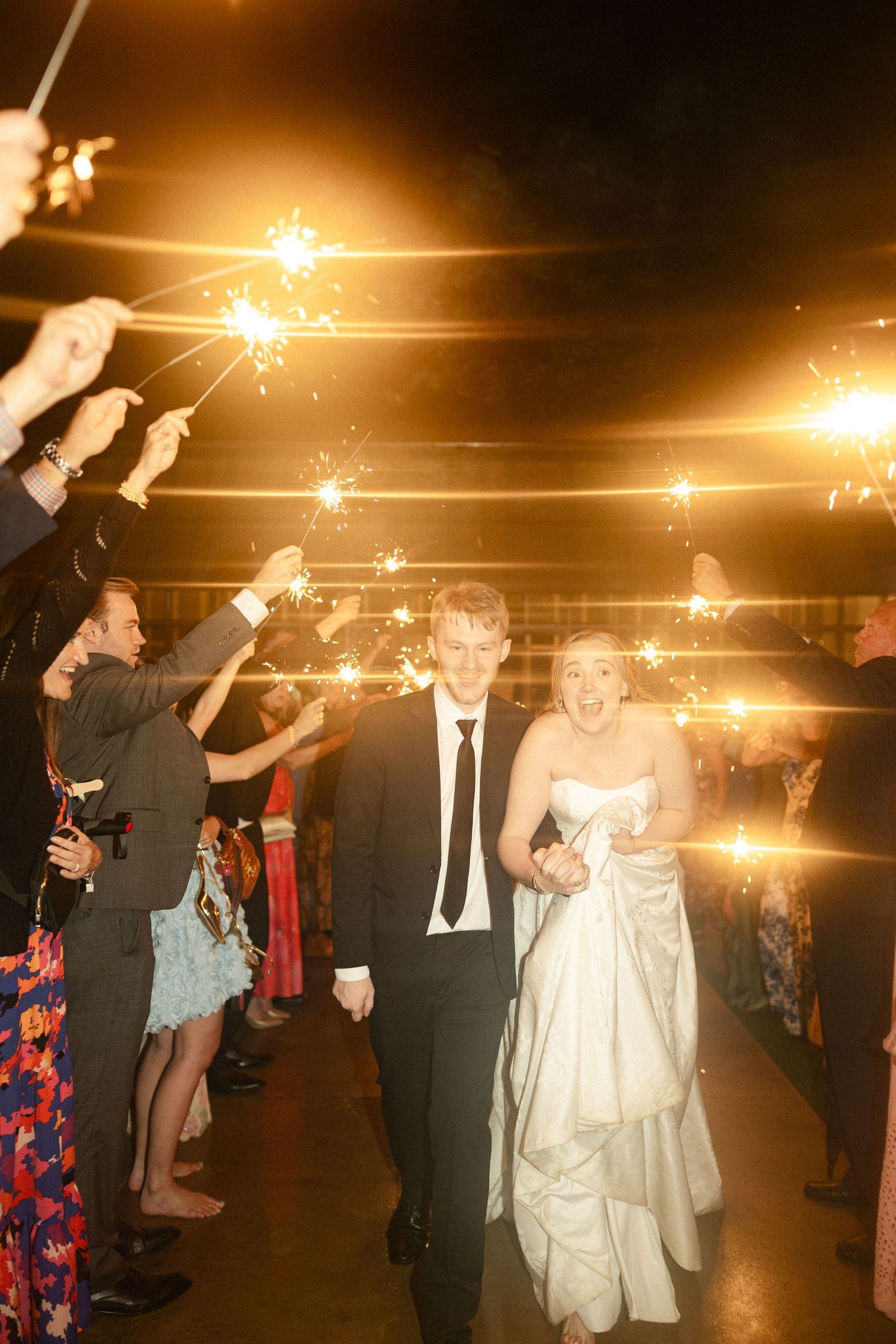 Newlyweds exit wedding reception through a sparkler tunnel surrounded by cheering guests at night.