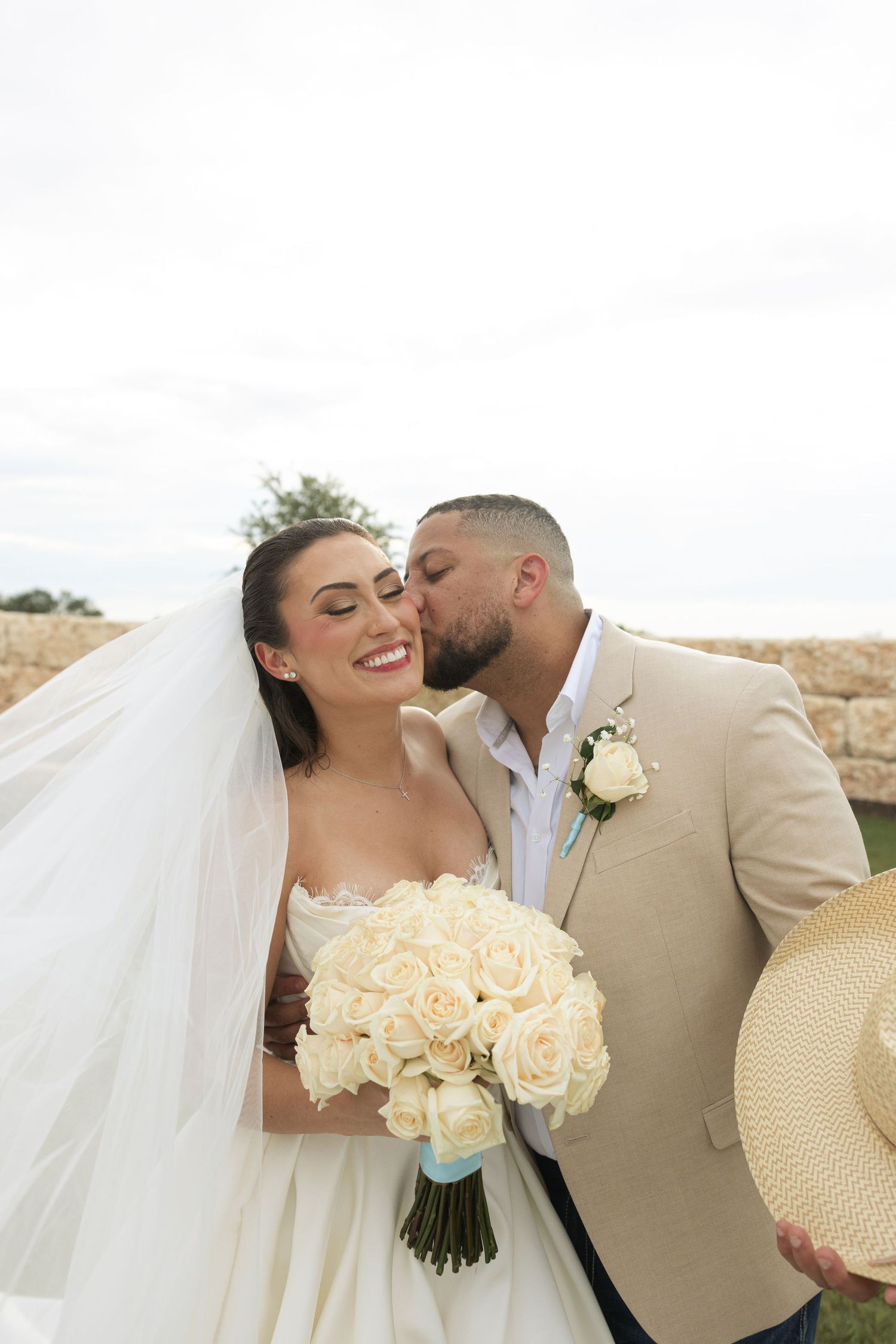 A romantic wedding couple in beige and white sharing intimate kisses while holding a bouquet of cream roses.