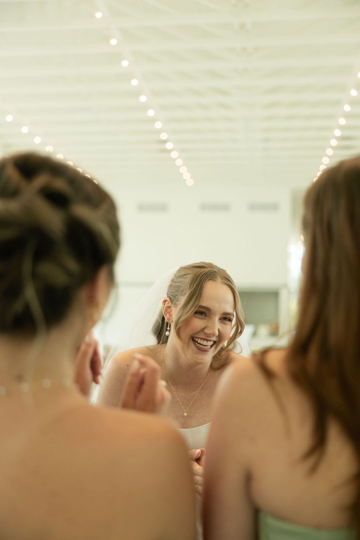 Smiling bride laughing with bridesmaids while getting ready in a bright venue with string lights.