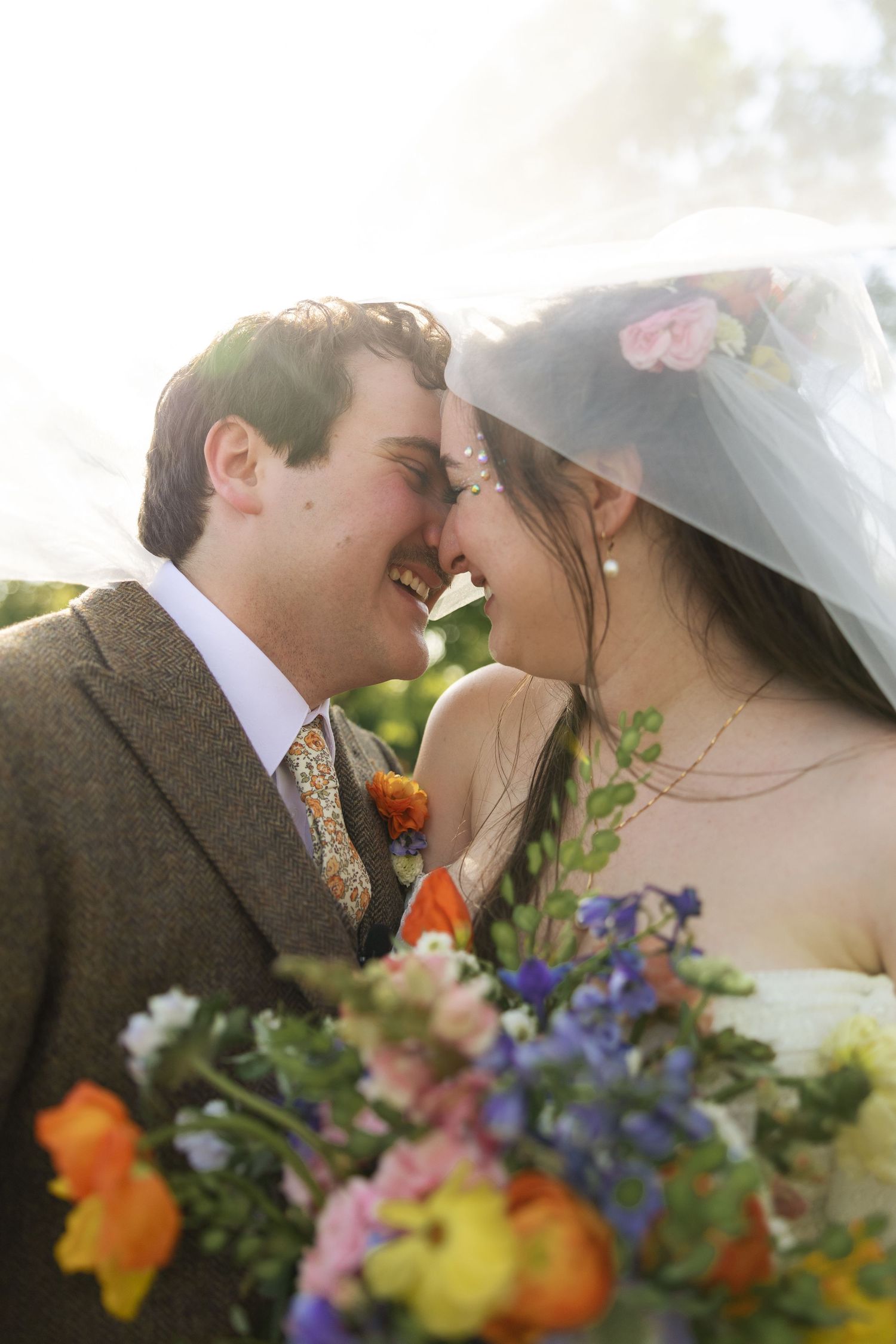 A bride and groom share a kiss behind a vibrant bouquet of colorful flowers.