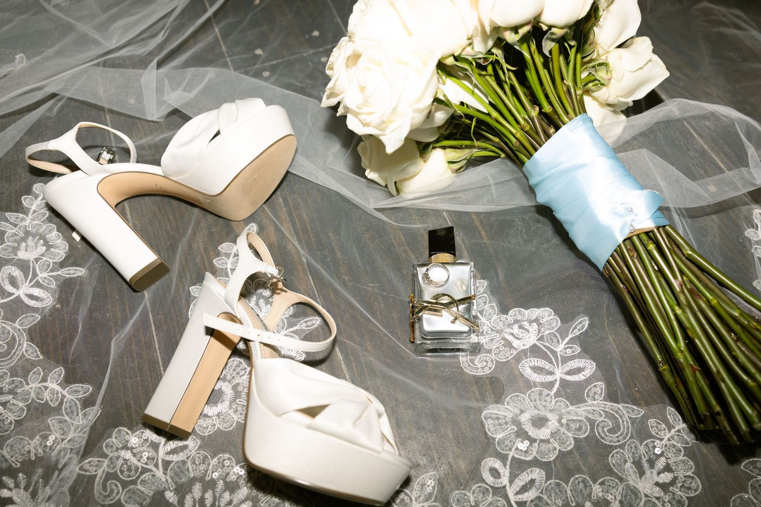Wedding accessories including white heels, a perfume bottle, and a bouquet wrapped in light blue ribbon.