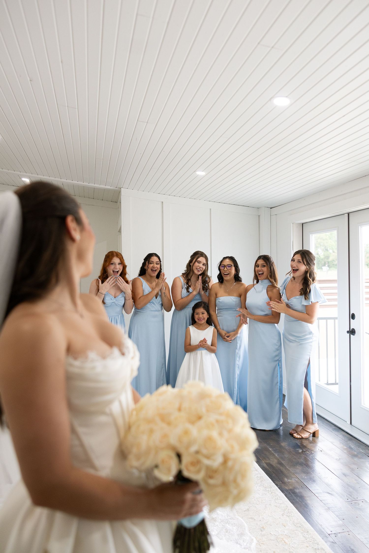 Bridesmaids in light blue dresses react emotionally to seeing the bride in her wedding gown.