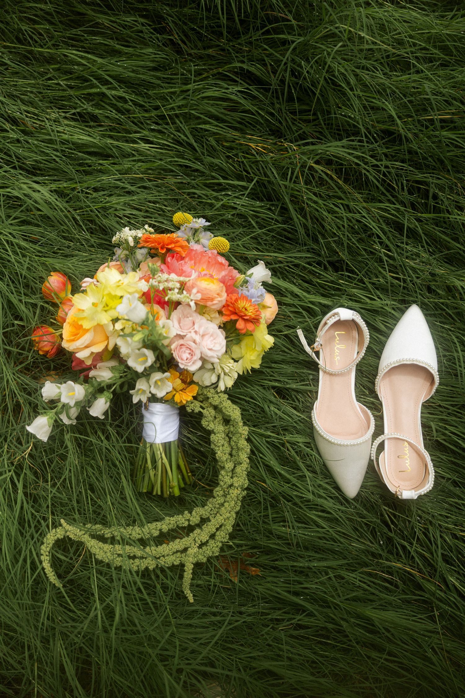 Colorful bridal bouquet with orange, pink, and yellow flowers beside white heels on lush green grass.