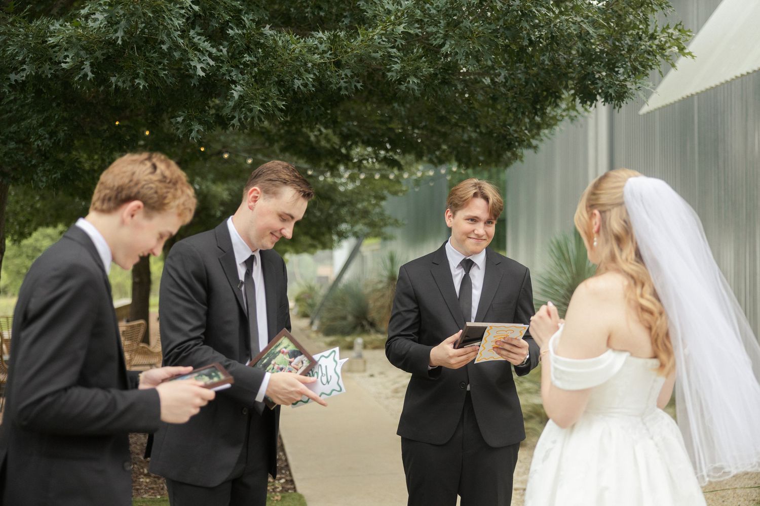 Bride in white gown and veil laughs with three groomsmen in black suits looking at phones outdoors.