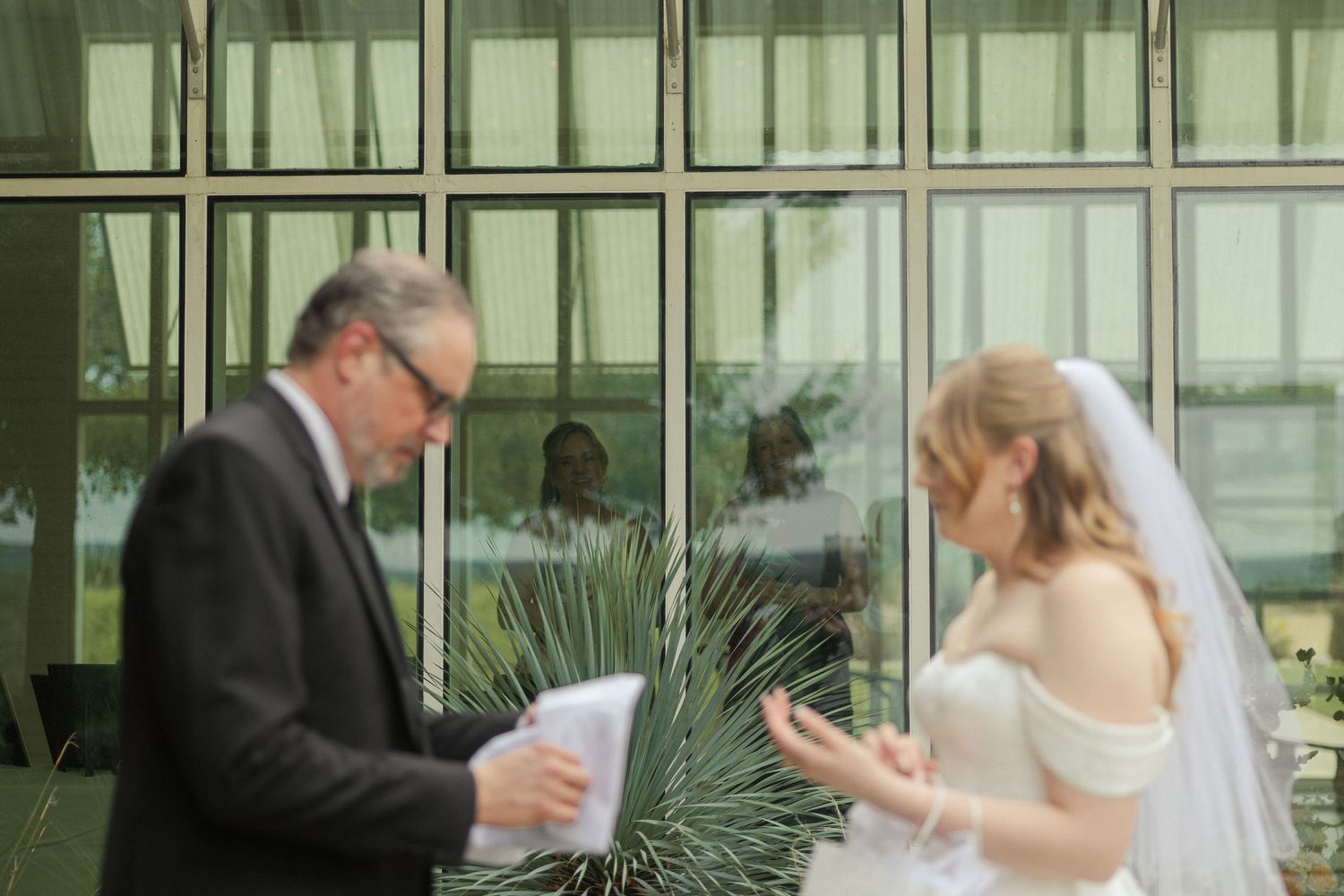 Bride in off-shoulder gown gestures while speaking with a man in a suit outdoors near glass windows.