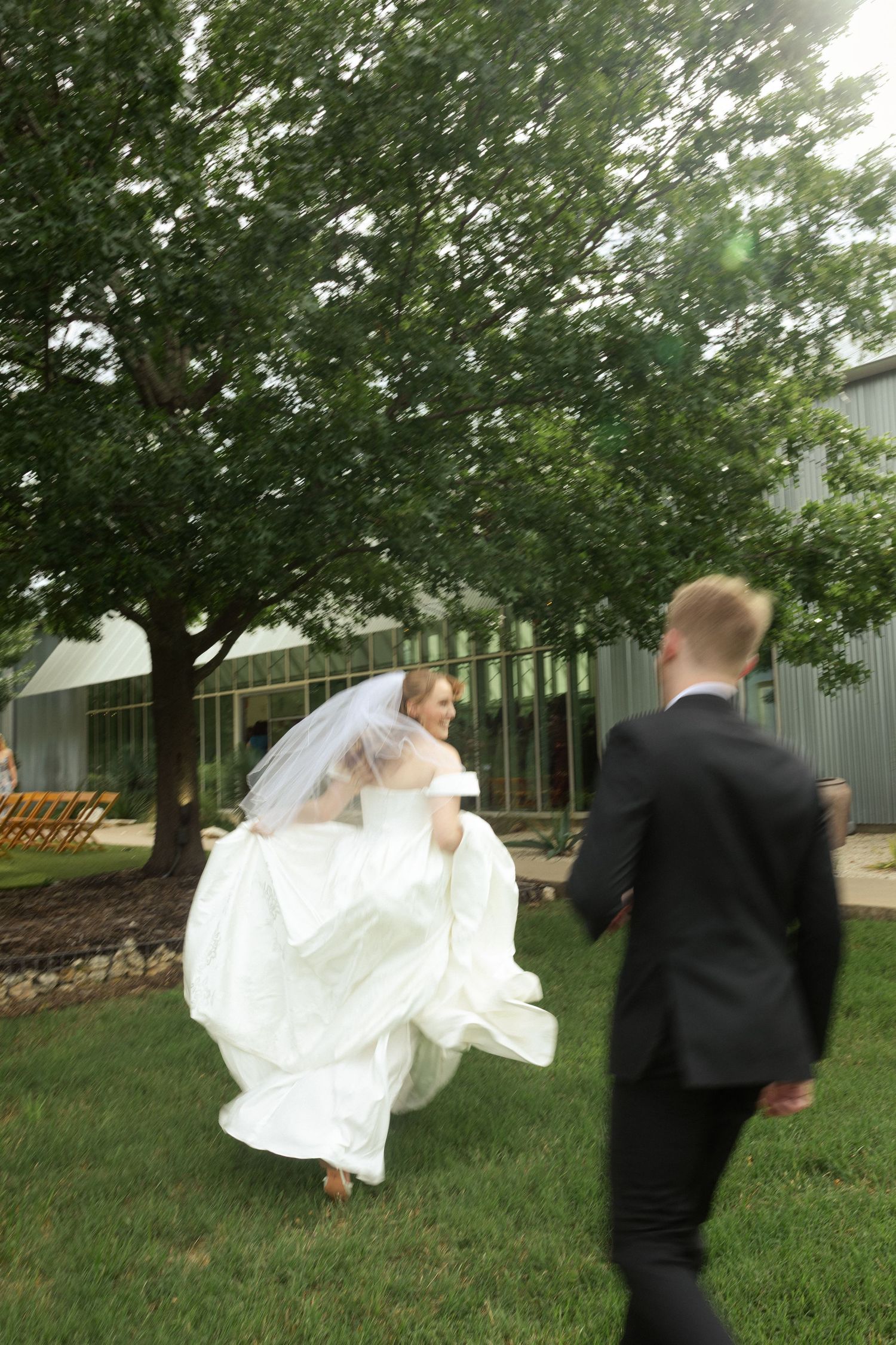 Bride in white gown and veil runs toward groom in black tuxedo on green lawn outside wedding venue.