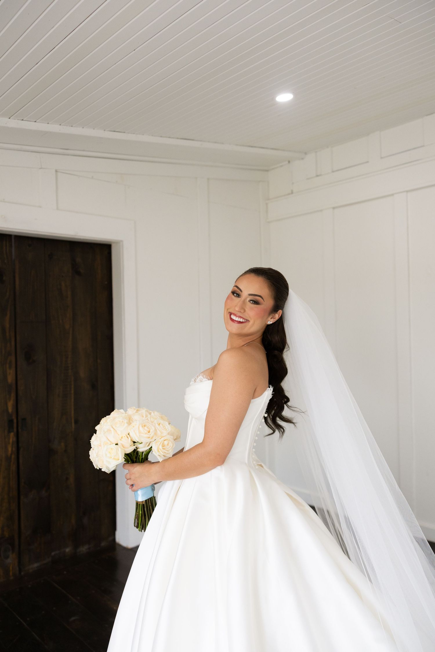 A bride wearing an elegant white ball gown holds a bouquet of white flowers against a light interior background.