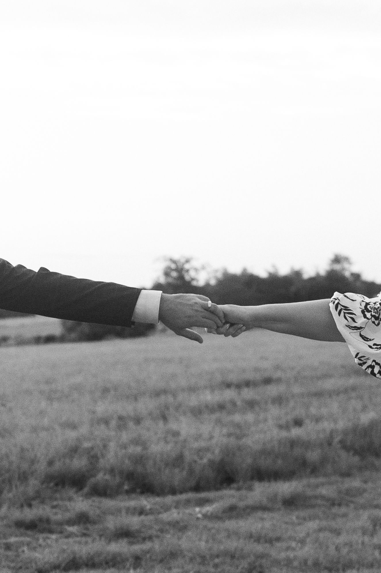 Black and white photo of couple holding hands across field landscape.