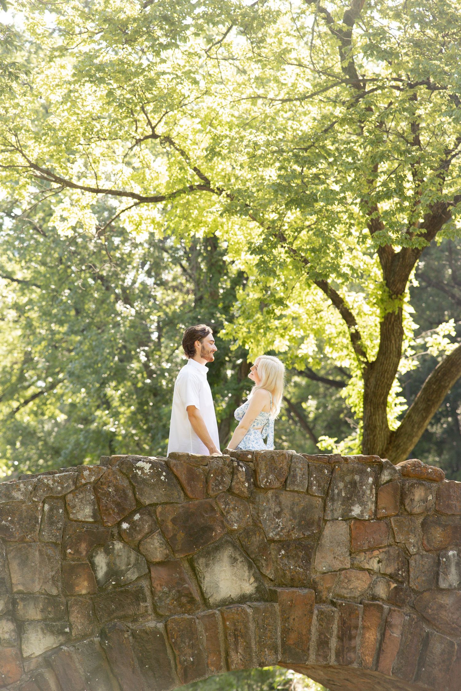 Couple sits atop rustic stone bridge wall surrounded by lush green trees.