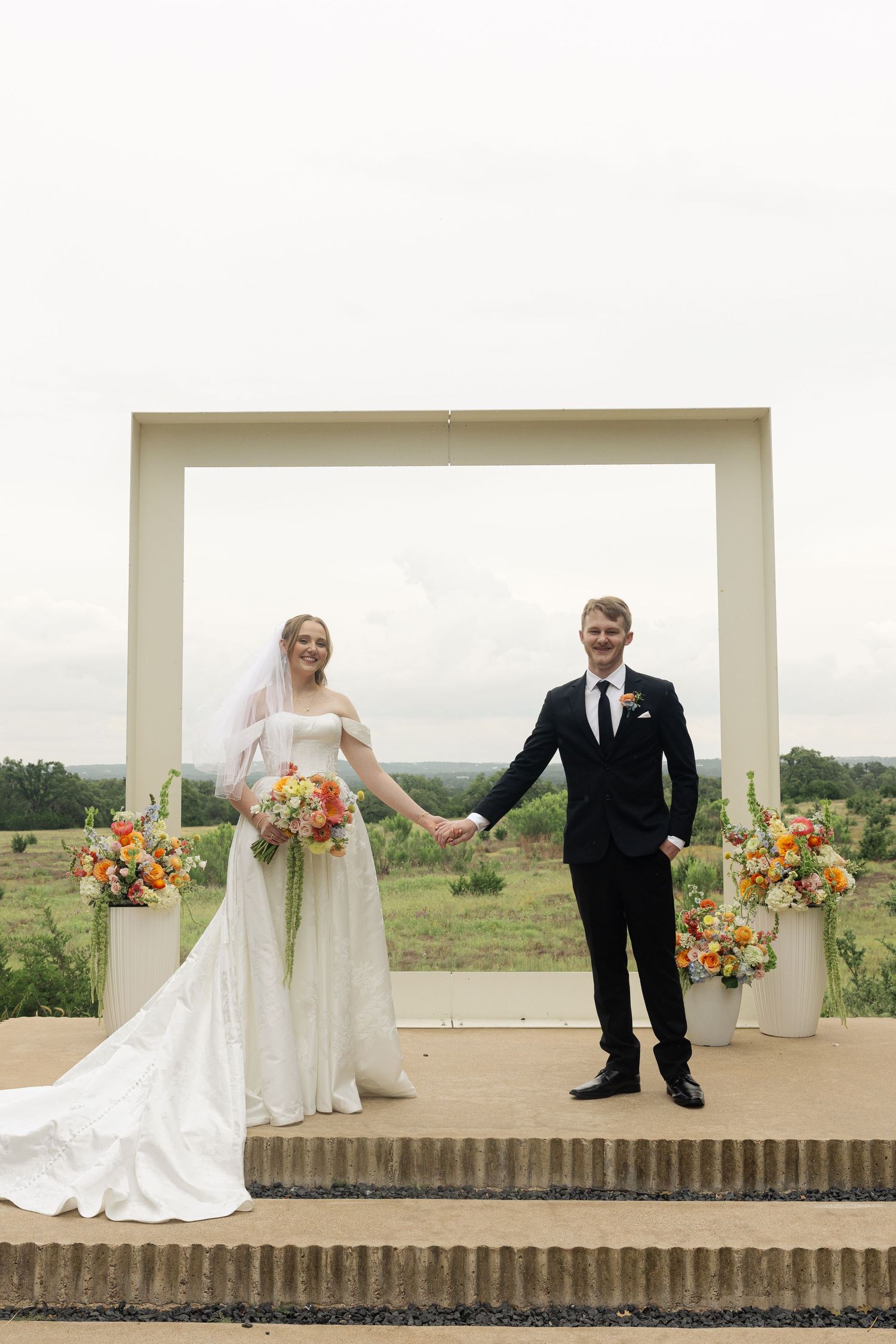 Bride and groom holding hands at outdoor altar with orange floral arrangements and scenic views.